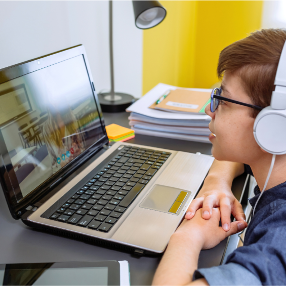 A young child sitting at a desk with headphones on, attentively watching a lesson on a laptop during an online class