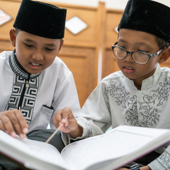 Two young Muslim children sitting side by side, reading from Qaida tul Quran with focus and guidance, in a bright learning environment.