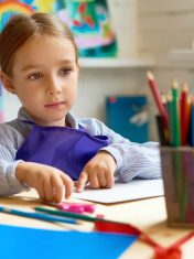 A cheerful little girl in a preschool classroom, smiling brightly while sitting at a table with colorful learning materials around her.